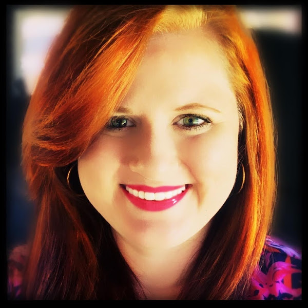 amanda-nevels-profile-pic A person with vibrant, flowing auburn hair styled in loose waves, wearing a floral patterned top, in a softly lit background.