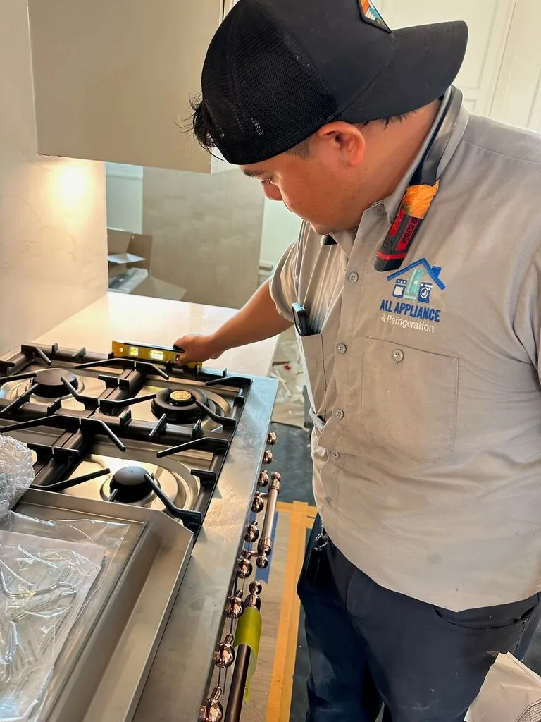 A technician wearing an 'All Appliance & Refrigeration' shirt is using a level to check the installation of a new stainless steel gas cooktop.