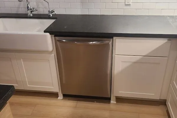 A stainless steel dishwasher built into a kitchen with white cabinets, a black countertop, and a white farmhouse sink.