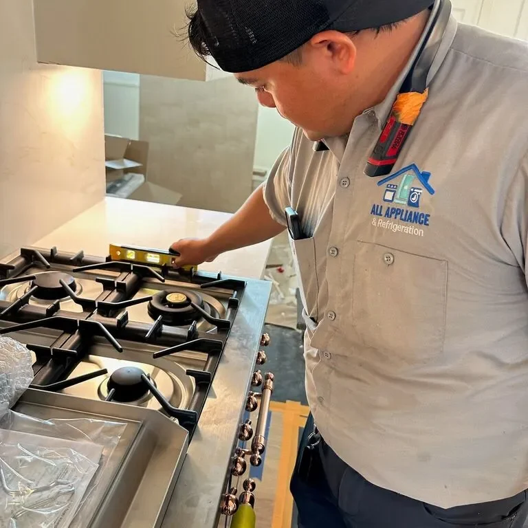 A technician wearing an 'All Appliance & Refrigeration' shirt is using a level to check the installation of a new stainless steel gas cooktop.